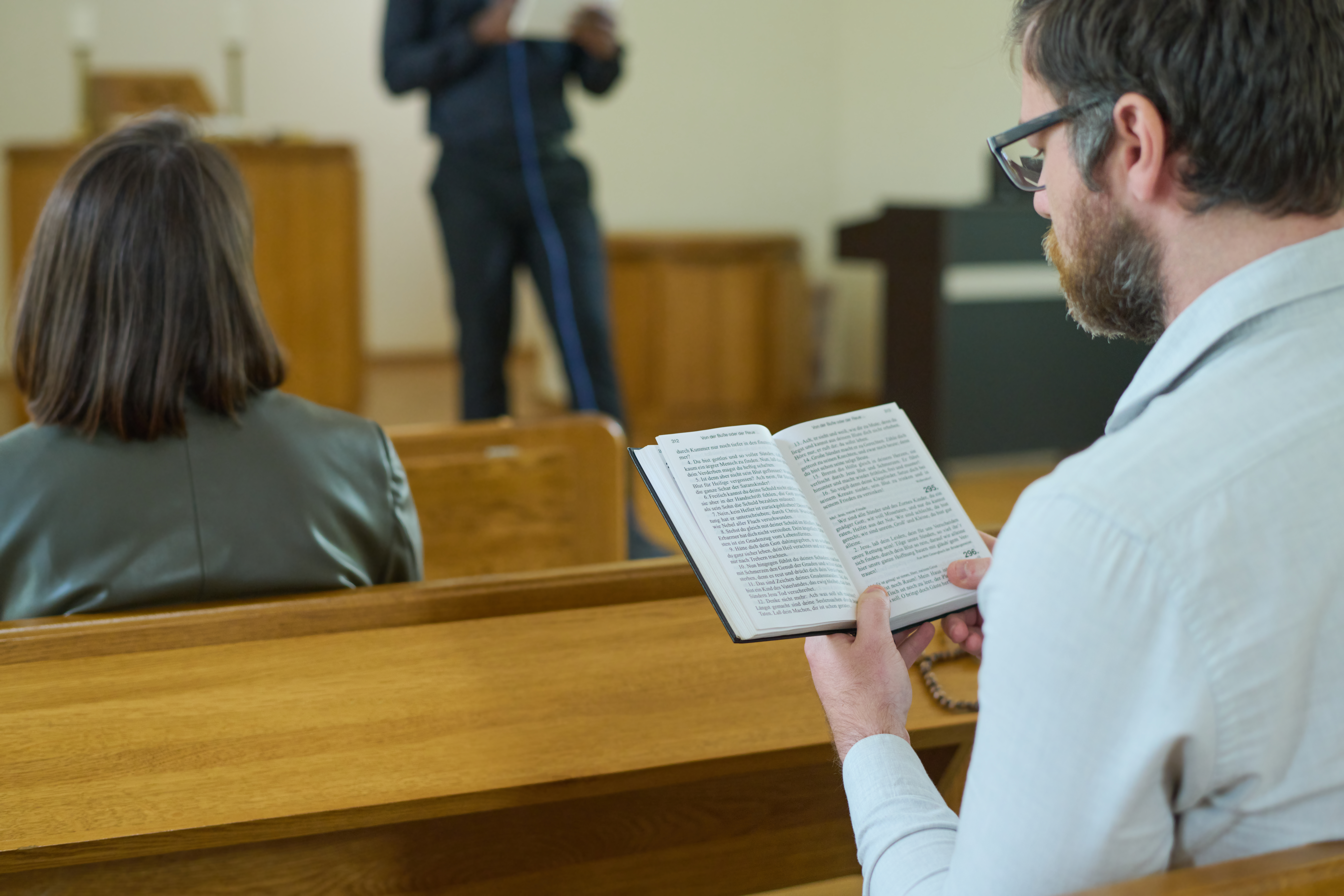 Middle aged man with open Holy Bible reading verses from Gospel while sitting on bench by desk and listening to sermon during church service